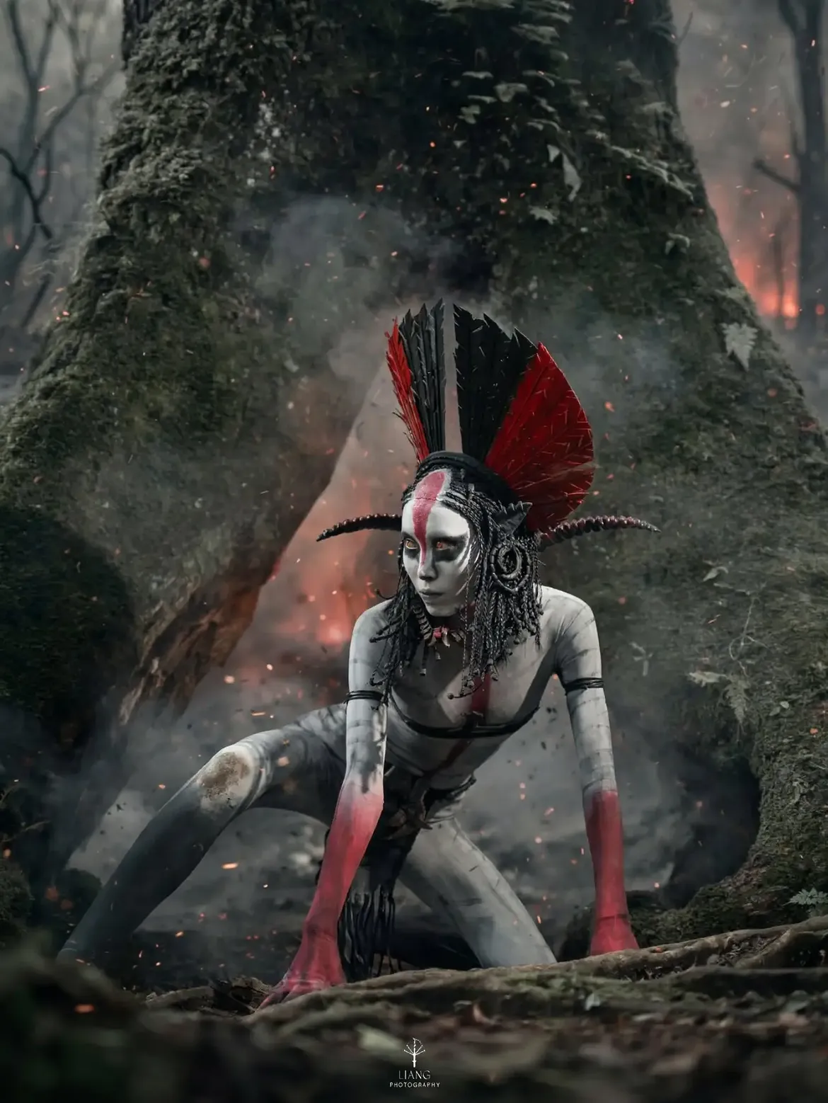 The Cosplay Monster in an original creature costume: white and red body paint, a black and red feathered headdress and dreadlocks, crouching in a smoke-filled forest.